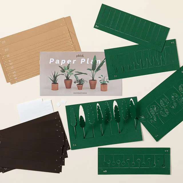 A flat lay display of an "Unkillable Desk Plants" paper DIY kit featuring an instruction card with potted plant illustrations, tan and dark brown lined paper sheets, and multiple green paper sheets with pre-cut plant and pot designs ready for assembly.