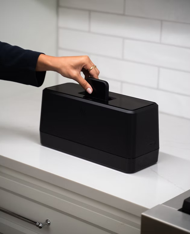 A person's hand reaches toward a sleek black rectangular device with a handle on top, sitting on a white surface against a white subway tile backdrop. The Swippitt Toaster Charger Thing for Your Phone is a compact black charging station with a modern, minimalist design.