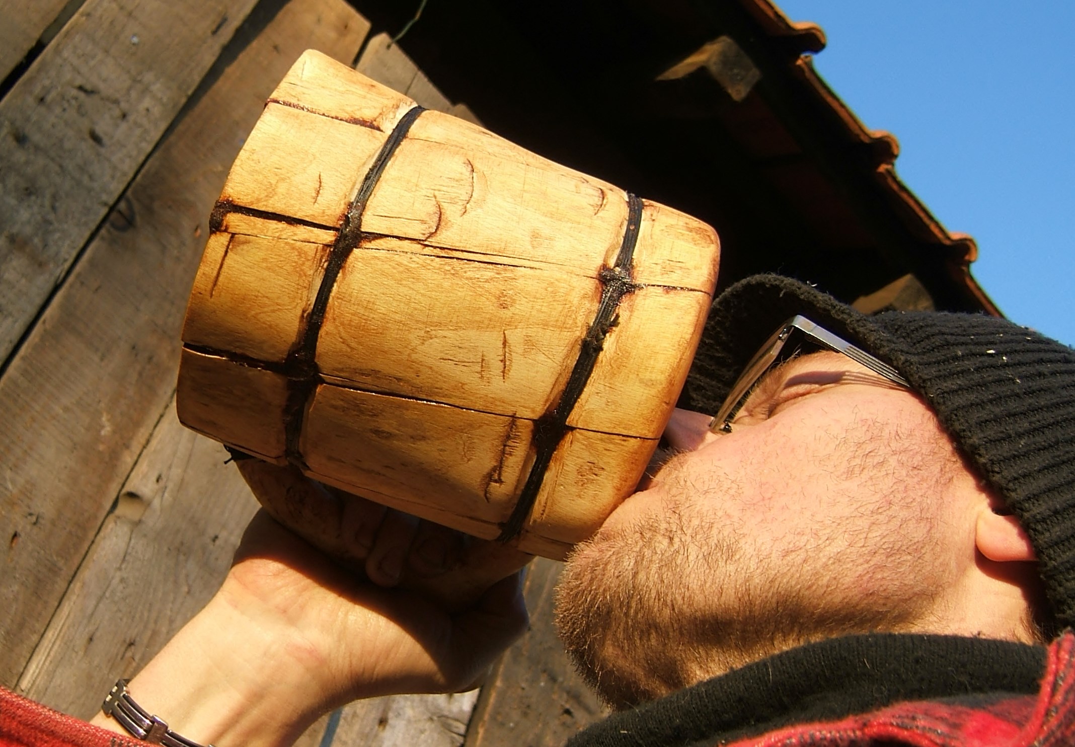 A wooden beer mug with a cylindrical body made from light-colored wood staves held together by dark leather straps, displayed in a man's hand against a wooden structure and blue sky background.