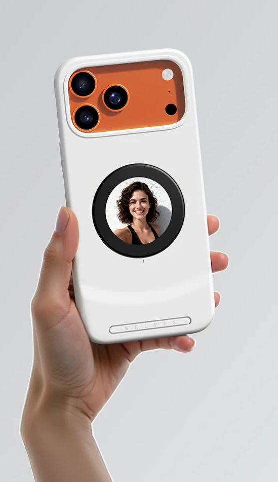 White Selfix iPhone case with orange camera module and a 1.5-inch rear circular display showing a portrait photo, held in a hand against a gray background.
