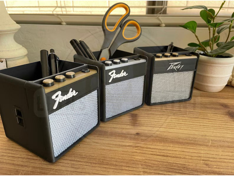 Three miniature black and gray Fender and Peavey guitar amplifier-shaped desk organizers with functional storage compartments, displayed on a wooden desk holding pens, scissors, and other office supplies next to a potted plant.