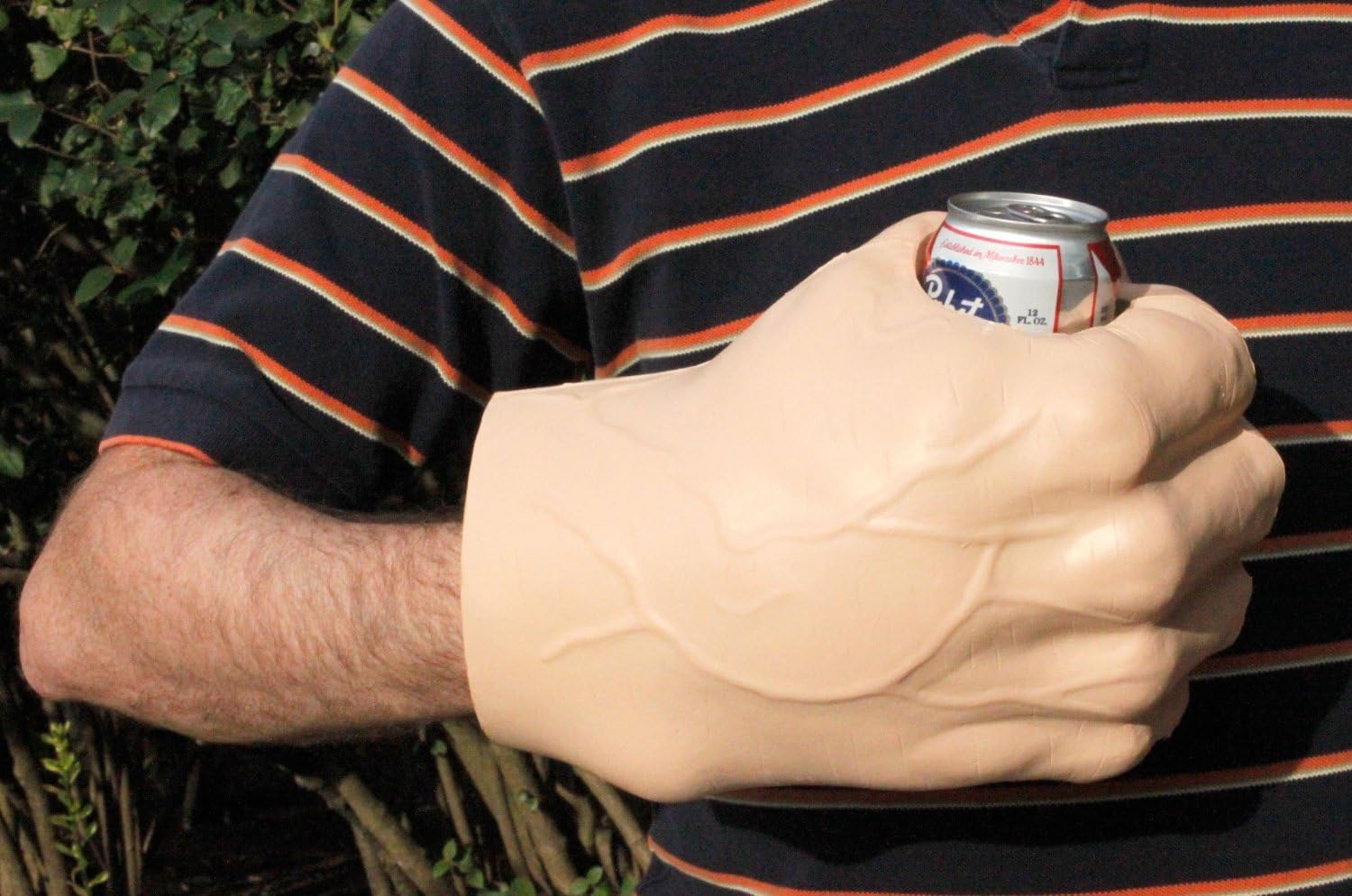 A large foam novelty hand in flesh tone gripping a beer can, worn over the arm like a glove, displayed against a striped shirt with green foliage in the background.