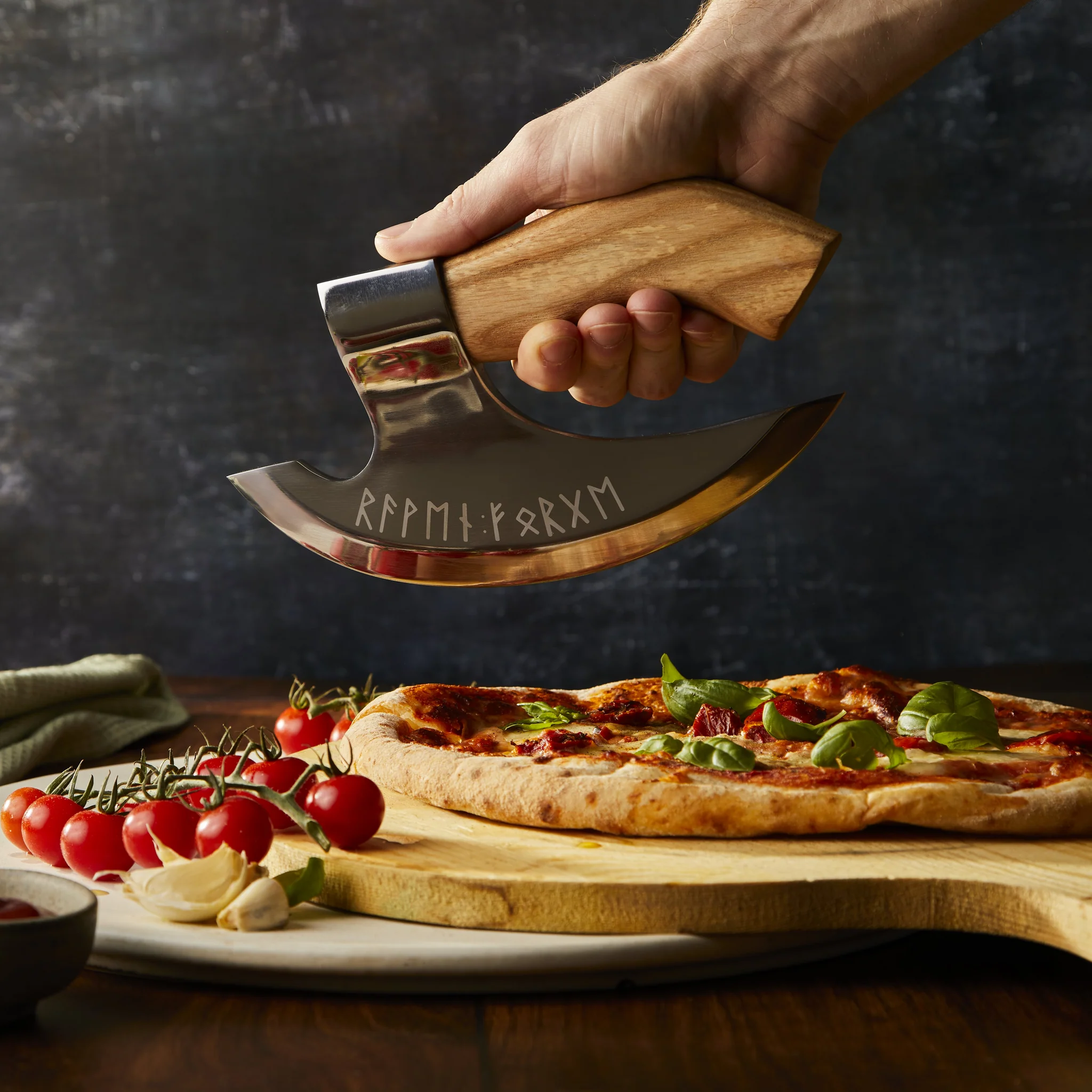 A curved stainless steel pizza axe with a wooden handle is being held above a freshly baked pizza surrounded by fresh tomatoes and herbs on a wooden board. The axe features engraved runic text along its curved blade and is designed for cutting and serving pizza in a distinctive Viking-inspired style.