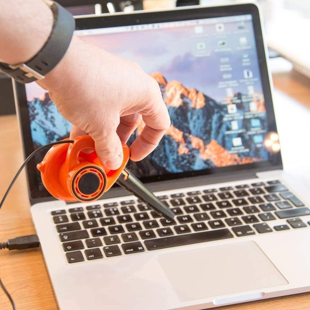 A bright orange and black miniature desk leafblower is held in someone's hand above a laptop keyboard, displaying its compact size and novelty design as a functional desktop accessory.