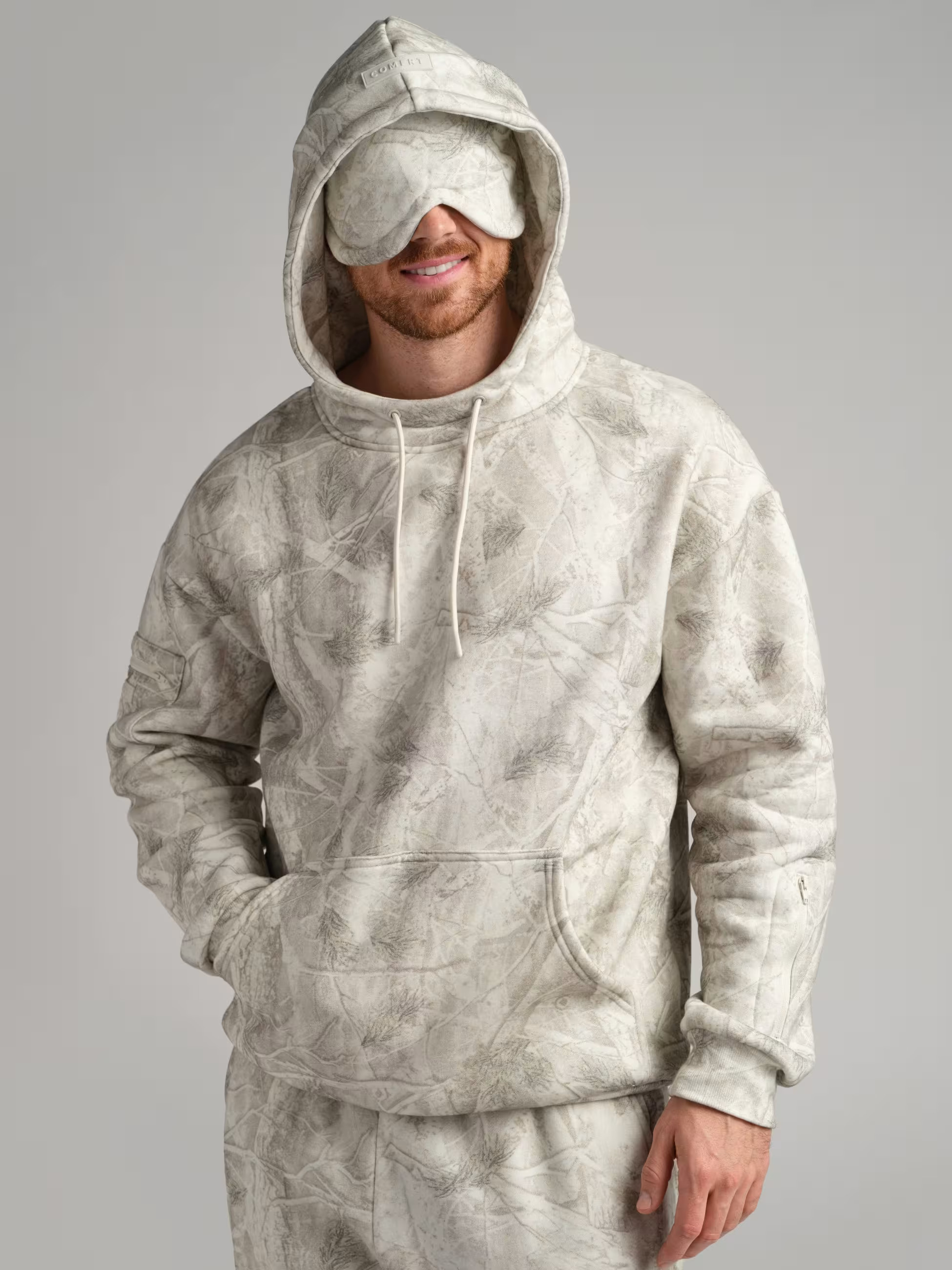 A cream-colored tie-dye patterned hoodie featuring an attached oversized eyemask that drapes over the wearer's eyes, paired with drawstring hood details and a kangaroo pocket for a cozy, all-in-one travel comfort outfit.