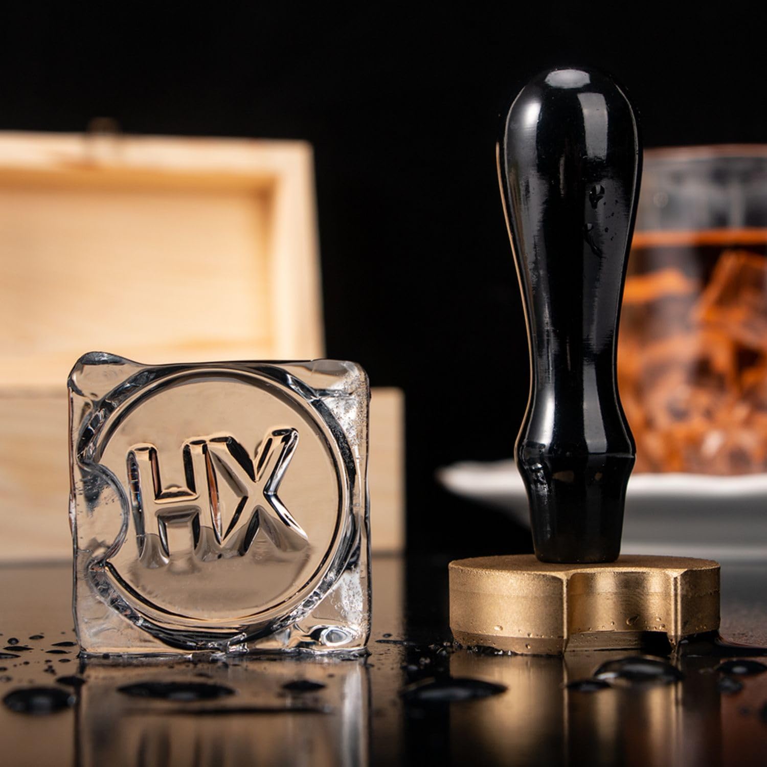 A customizable ice cube stamp featuring a clear square mold with "HX" engraved in the center, accompanied by a black handle with a gold-colored base, displayed on a dark surface with scattered decorative elements.