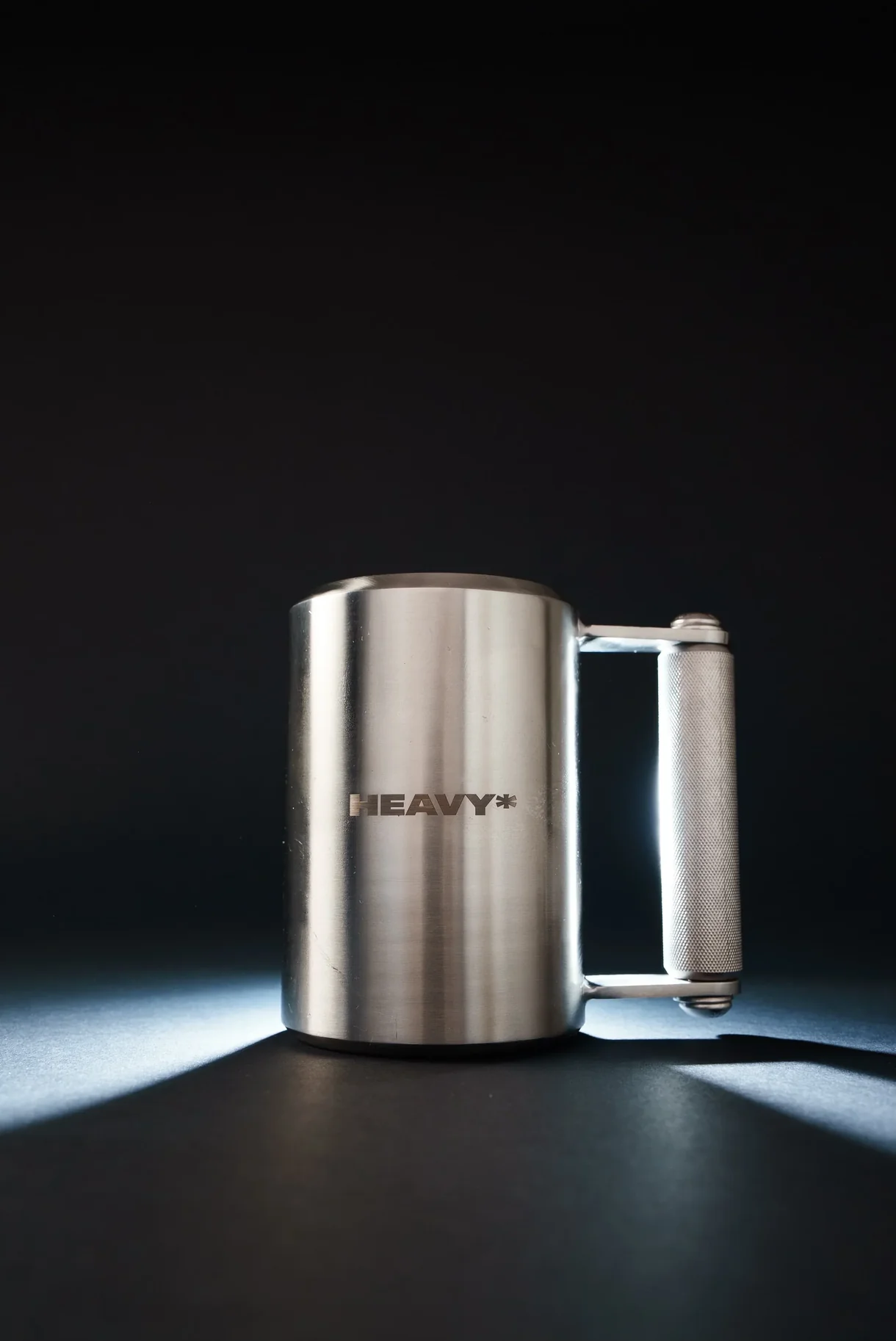 A sleek stainless steel mug branded "HEAVY*" featuring a polished cylindrical body and a textured black handle, photographed against a dark background with dramatic lighting.