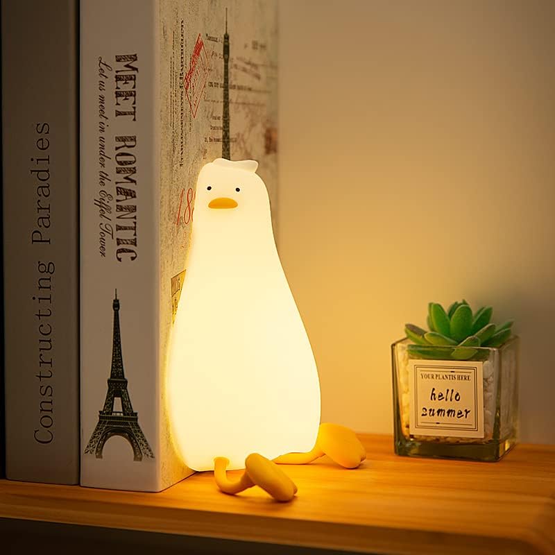A soft-glowing silicone duck-shaped night light with a minimalist design, featuring a simple face with dot eyes and orange beak, emitting warm ambient light on a wooden shelf alongside books and a succulent plant.